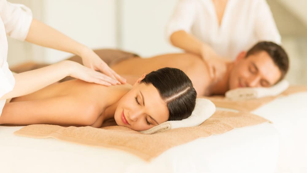 Two people enjoy a couples massage, lying side by side on tables as therapists provide soothing back massages in a calm, spa-like setting. Both appear relaxed with eyes closed. For more details, check our relaxation massage FAQ.