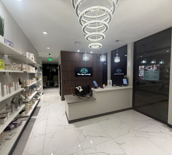 Reception area of a modern spa with lighted chandeliers, a marble floor, product displays on shelves, and two computer stations at the front desk.