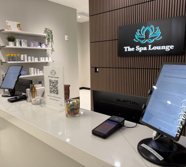 Reception desk at The Spa Lounge with two screens, a QR code sign, business cards, a candy jar, and beauty products on shelves in the background.