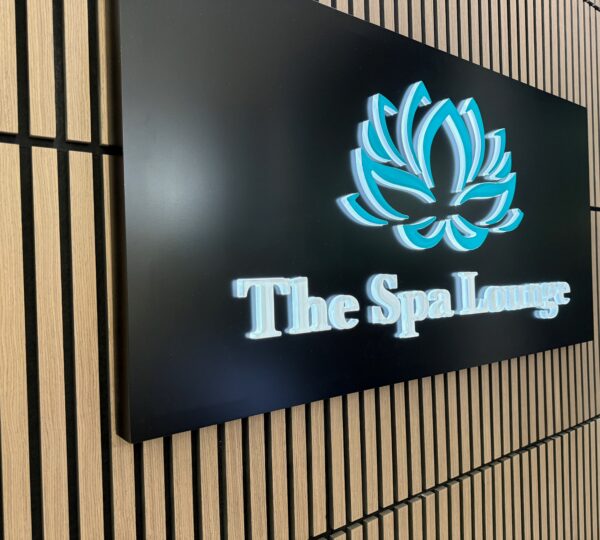 A black sign with a blue lotus flower logo and the words "The Spa Lounge" mounted on a light wooden slat wall.
