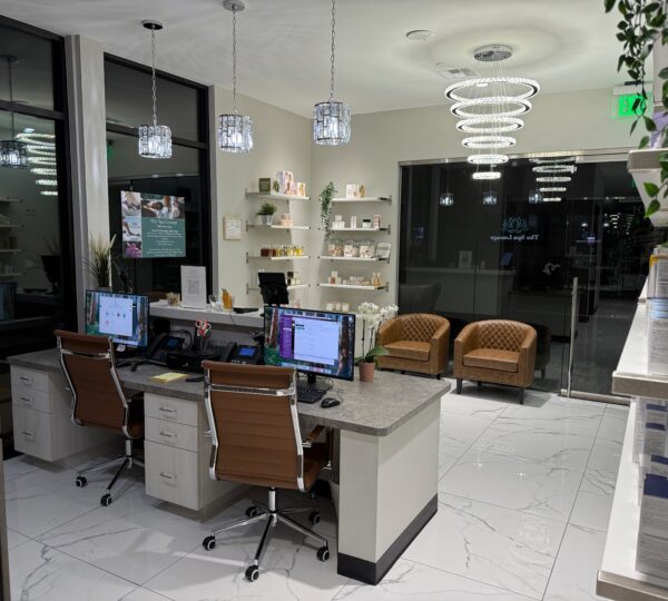 A modern reception area with two desks, computers, brown chairs, shelves with products, large windows, and white tile flooring.