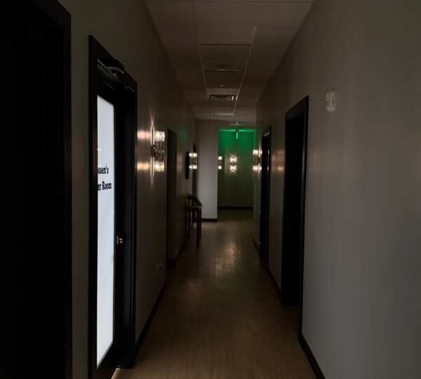 Dimly lit hallway with closed doors on both sides, illuminated exit sign at the far end, and faint light coming from one room on the left.