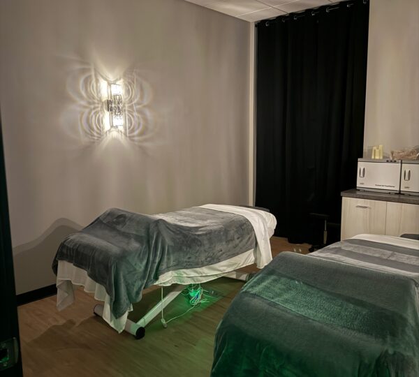 A dimly lit massage room features two massage tables with gray blankets, a wall sconce, black curtains, and cabinetry along the back wall.