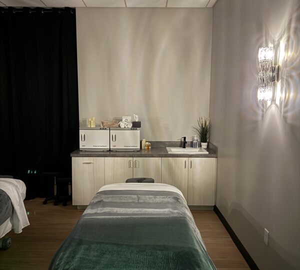 A massage therapy room with a treatment table, a countertop with supplies, cabinets, a plant, and a wall-mounted light fixture.