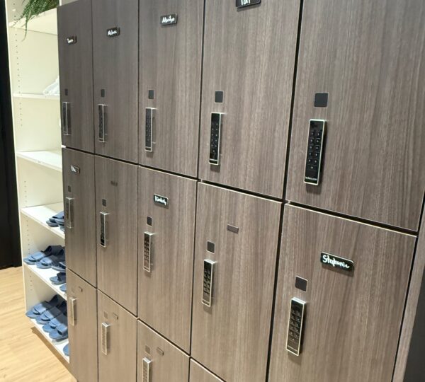 A set of twelve wood-finished lockers with digital locks, each labeled with a name, stands next to white shelves holding folded clothing and slippers.