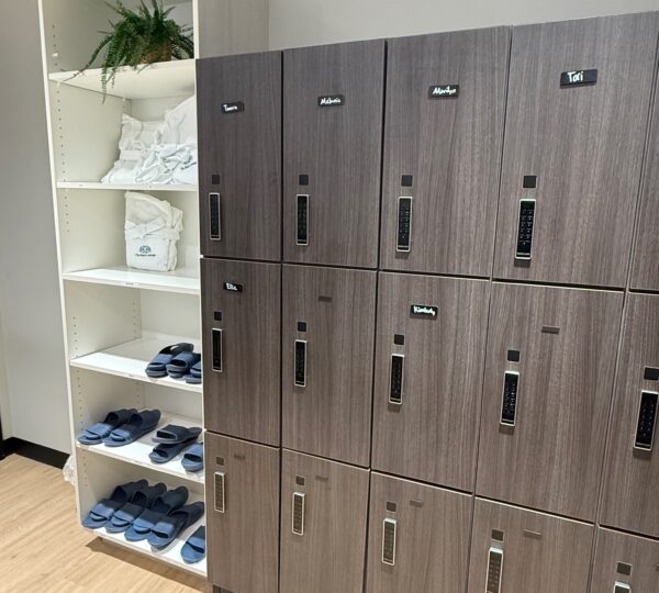 A set of gray lockers stands next to white shelves holding neatly folded towels, slippers, and a potted plant on the top shelf.