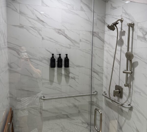 Accessible walk-in shower with marble tile, a glass door, grab bars, handheld showerhead, and three black wall-mounted dispensers.
