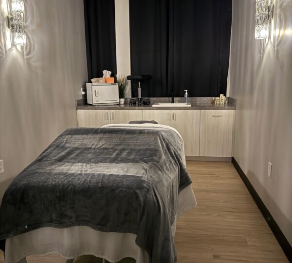 A massage or treatment room with a bed covered in gray and white linens, cabinets, countertop, and two wall lights. Black curtains cover the window at the back.