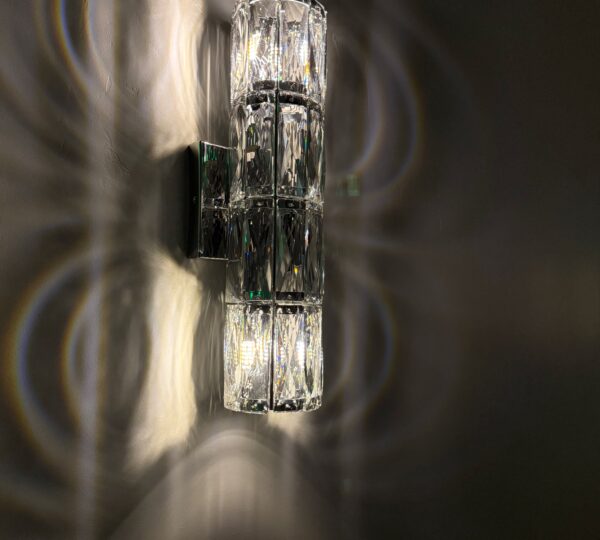A modern wall sconce with a geometric glass design casts patterned light and shadows on a gray wall.