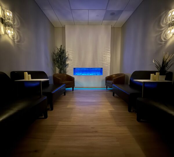 A dimly lit lounge with black sofas, wicker chairs, small tables with candles, potted plants, and a blue-lit wall feature at the center.