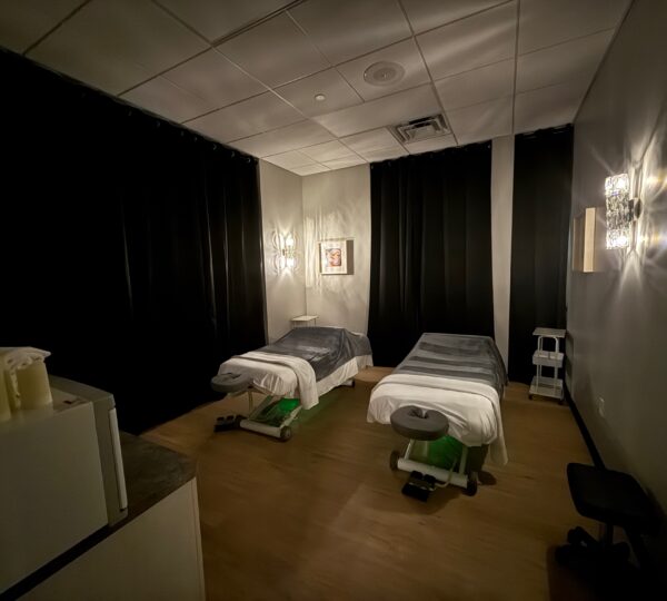 Dimly lit massage room with two massage tables, black curtains, wall art, and soft lighting, creating a calm and quiet atmosphere.