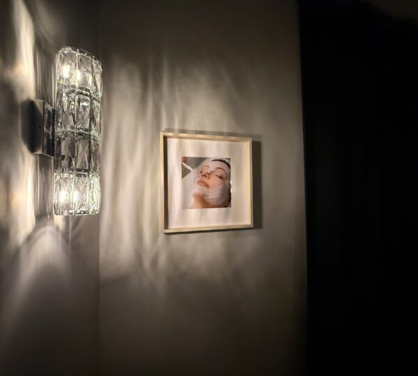 Wall-mounted light fixture illuminating a framed photo of a person with a towel wrapped around their head, hanging on a light-colored wall next to a dark curtain.
