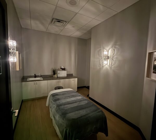A dimly lit massage room with a single massage table, gray blanket, wall lights, and a countertop with cabinets and a towel warmer.