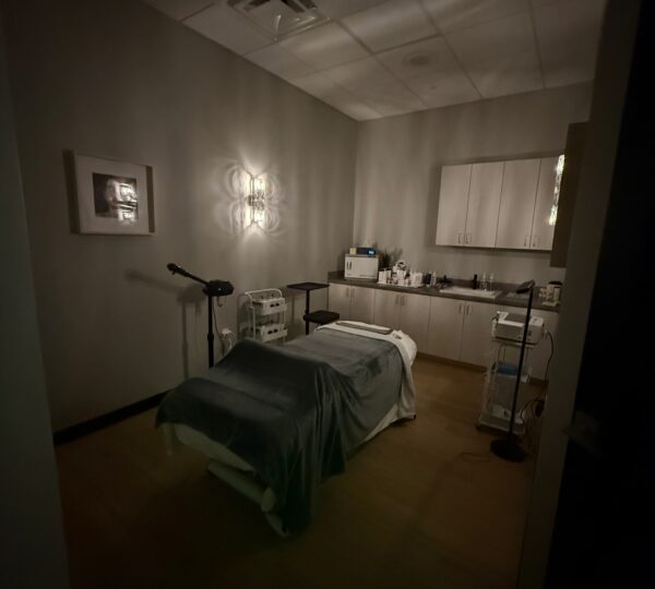 A dimly lit room with a massage table covered by a gray blanket, cabinets, medical equipment, and a wall-mounted lamp.