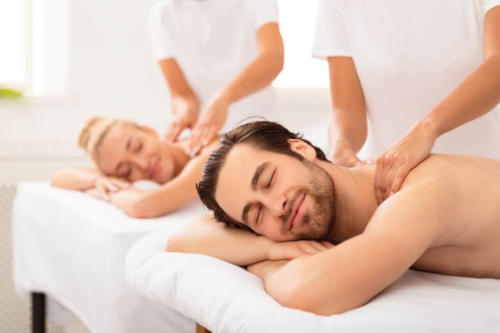A man and a woman receiving a couples deep tissue massage from two therapists while lying on spa tables.