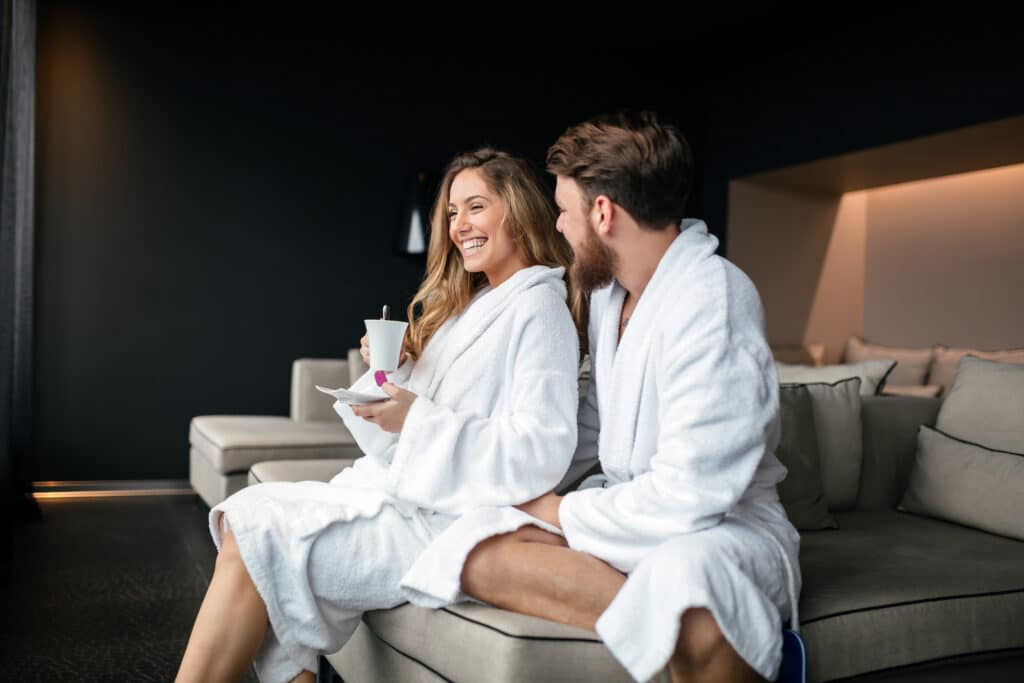 A man and woman in white bathrobes sit on a sofa, smiling at each other during a romantic spa experience.
