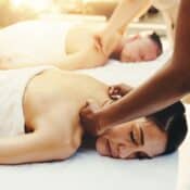 A massage therapist provides a deep tissue massage to a woman on a spa table, with another person receiving a couples massage in the background.