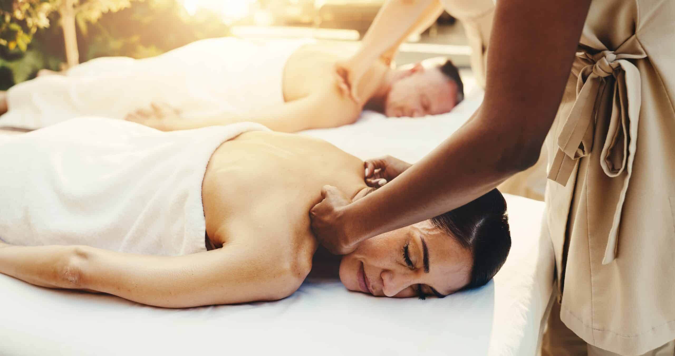 A massage therapist provides a deep tissue massage to a woman on a spa table, with another person receiving a couples massage in the background.