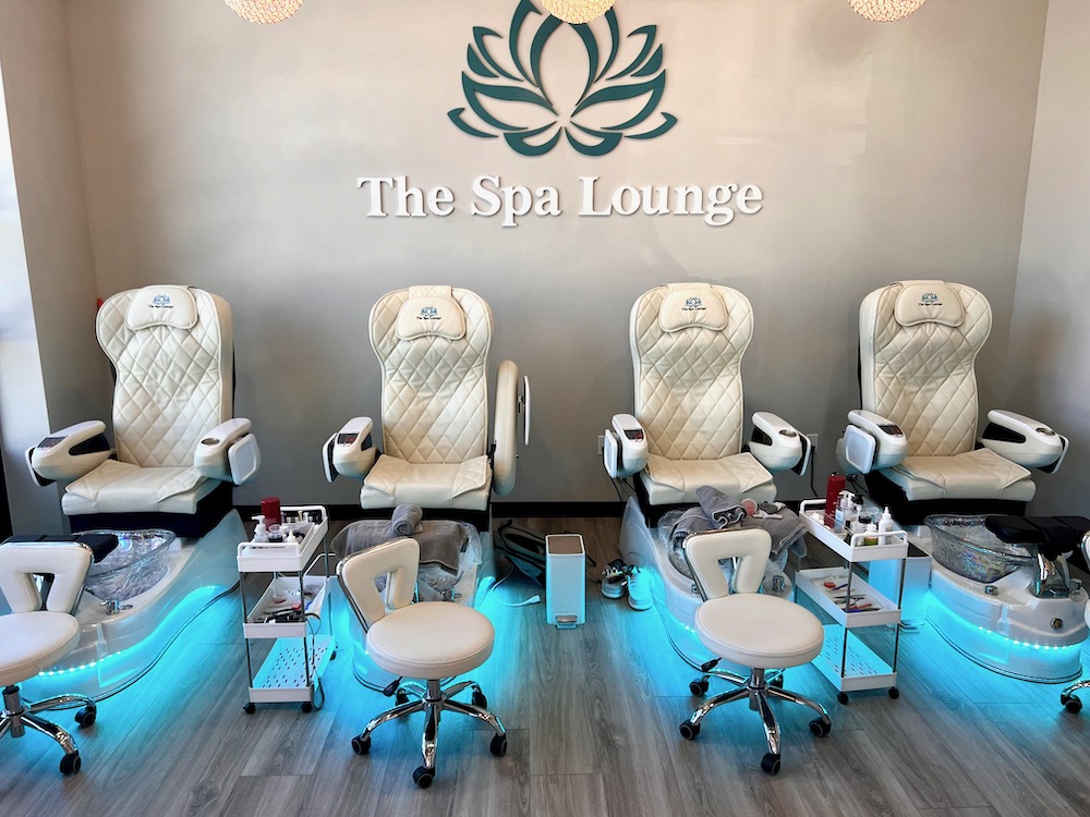 Four white Spa Lounge Pedicure chairs with foot baths and rolling stools are arranged in a row under a "The Spa Lounge" sign in a modern, well-lit spa.